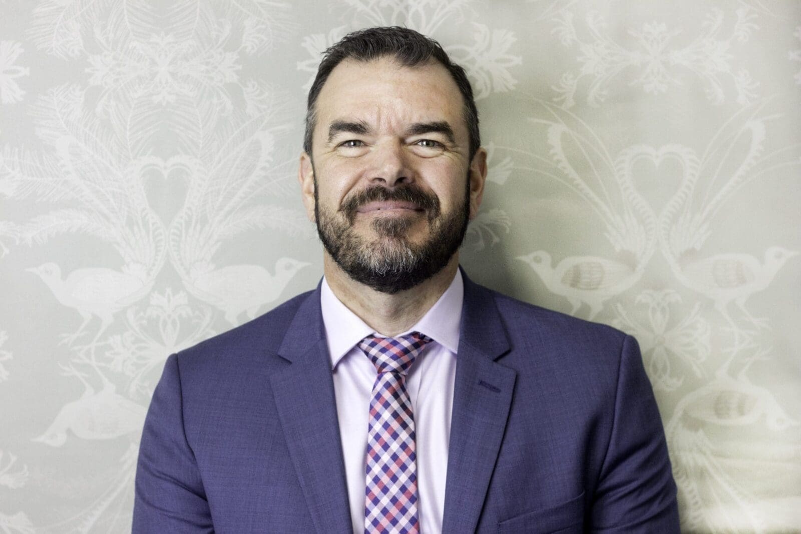 Man with beard wearing purple suit and checkered tie standing against patterned wallpaper.
