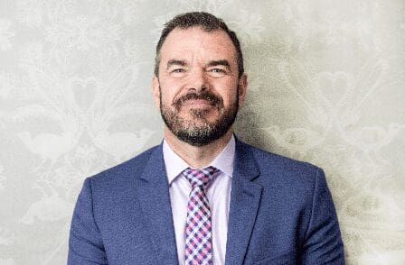 Man in blue suit jacket and patterned tie standing against a light floral and bird patterned wall.