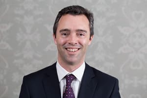 Man in dark suit, white shirt, and patterned tie smiling against grey patterned background
