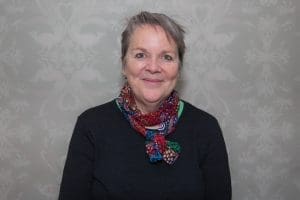 Middle-aged woman with grey hair tied back wearing a black top and colourful scarf against patterned grey background