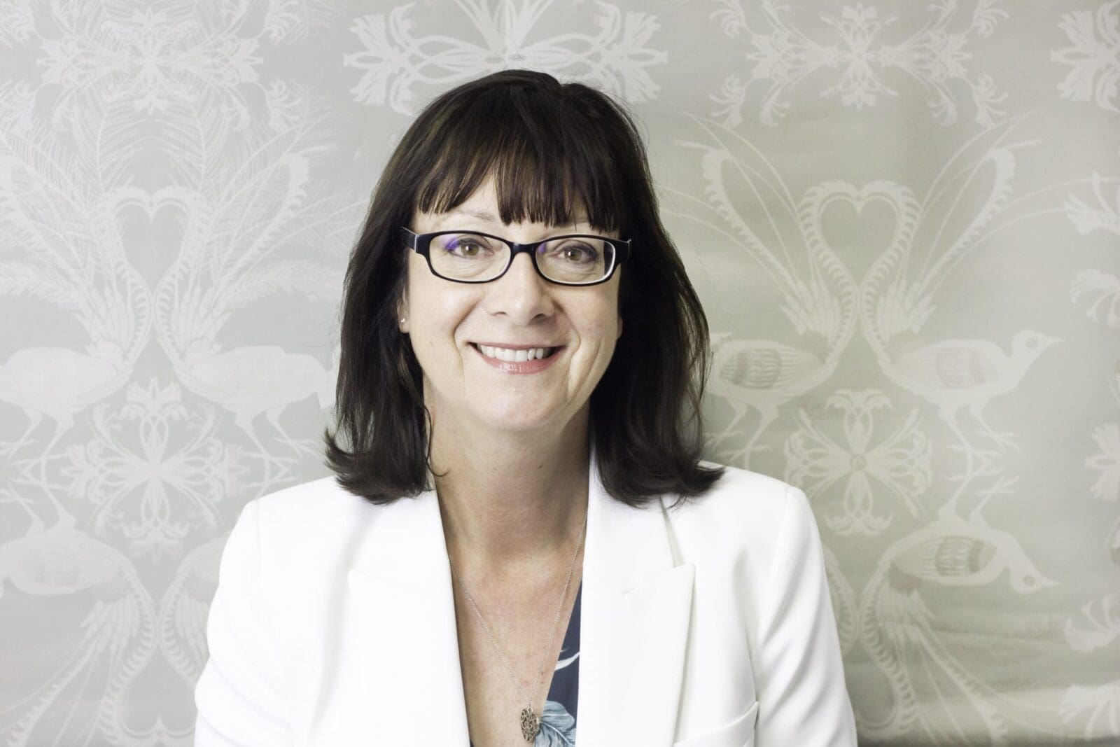 Middle-aged woman with glasses wearing white blazer and pendant necklace smiling against patterned wallpaper.