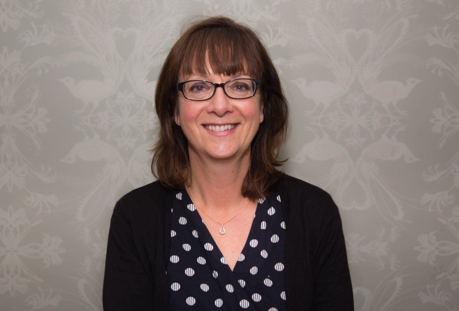 Woman with brown hair and glasses wearing a black cardigan over a navy polka dot top against grey wallpaper.
