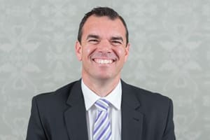 Man in dark suit and striped tie smiling against a light patterned background