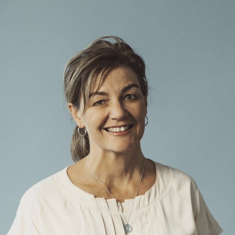 Middle-aged woman with light brown hair in ponytail, hoop earrings, light top, and pendant necklace.