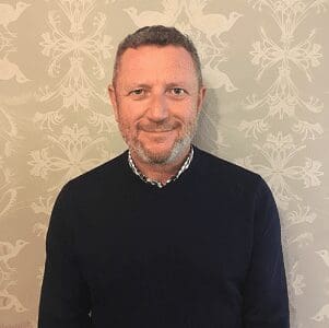 Middle-aged man with short curly hair and beard wearing dark sweater against patterned wall.