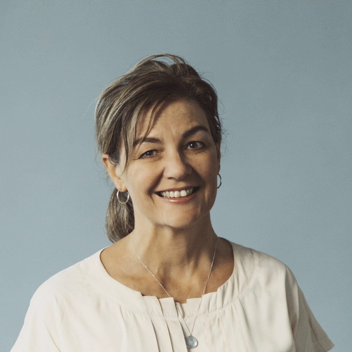 Middle-aged woman with light brown hair in ponytail, hoop earrings, light top, and pendant necklace.