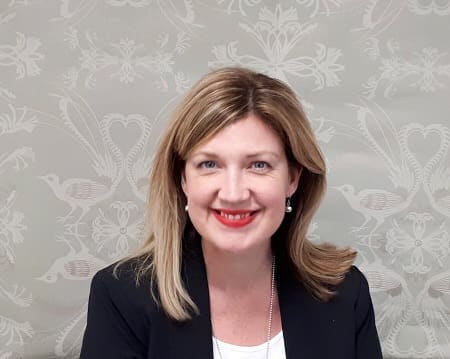 Smiling woman with shoulder-length blonde hair wearing black blazer and red lipstick against patterned background.