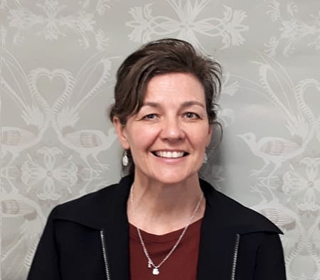Woman with short dark hair smiling, wearing black jacket and maroon top, standing by patterned wall