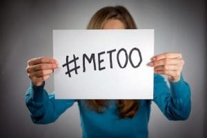 Person in blue shirt holding white paper with hashtag METOO written in black marker.
