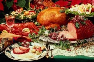 Festive table with roasted turkey, ham garnished with grapes, cooked lobster, and side dishes.