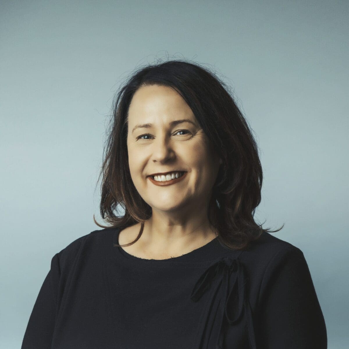 Portrait of a smiling middle-aged woman with shoulder-length dark hair wearing a black top against grey background