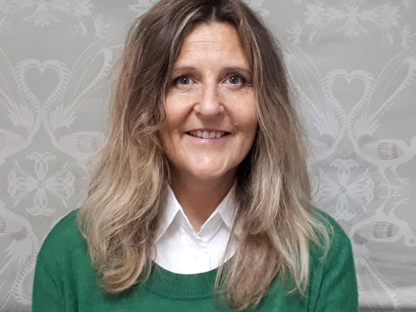 Woman with shoulder-length light brown hair wearing white collared shirt and green sweater against patterned background