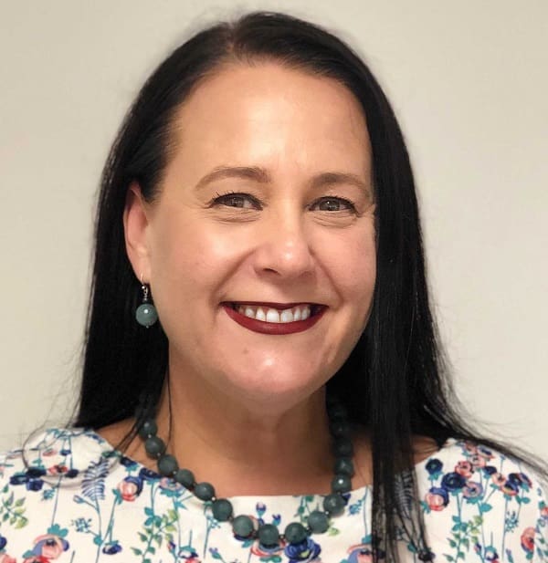 Woman with long dark hair wearing a floral top, dark lipstick, green bead earrings, and necklace smiling.