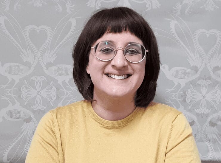 Person with short dark hair and round glasses wearing a mustard yellow top in front of patterned wallpaper