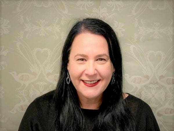 Woman with long black hair, black top, dangle earrings, smiling against a patterned background with birds and hearts