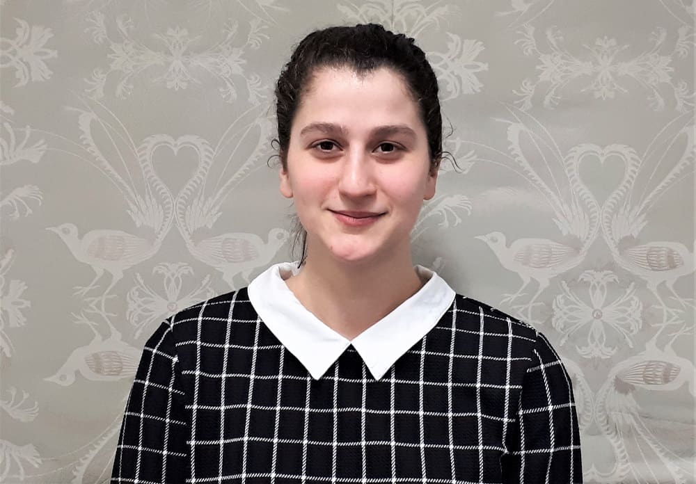 Young person with dark hair tied back wearing black and white checkered top and white collar, patterned wallpaper background.