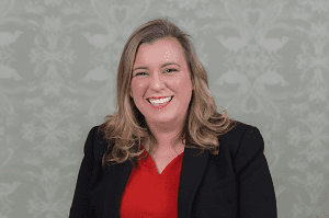 Woman with shoulder-length blonde hair wearing black blazer and red top against patterned background