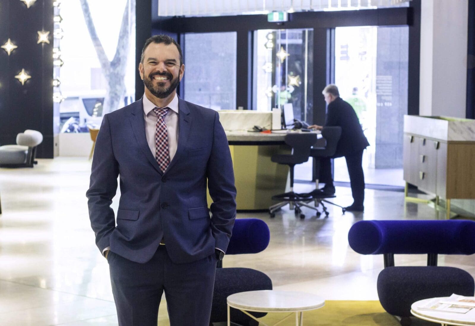 Man in blue suit standing in modern office lobby with person working at standing desk in background
