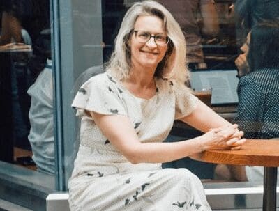 Woman with glasses sitting at a wooden table in a cafe wearing a light patterned dress.