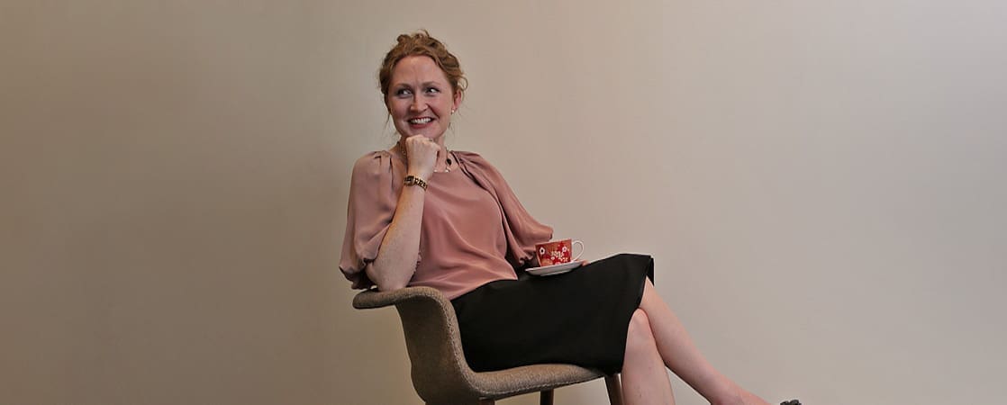Woman with red hair in pink blouse and black skirt sitting on chair holding teacup and saucer.