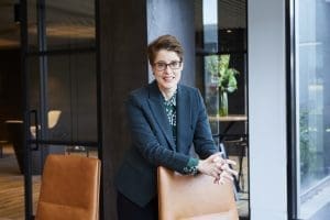 Person wearing glasses and dark blazer leaning on a brown leather chair in an office with glass walls.