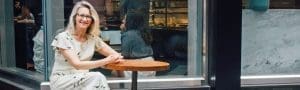 Woman with glasses sitting at a round wooden table outside a cafe with a glass window behind.