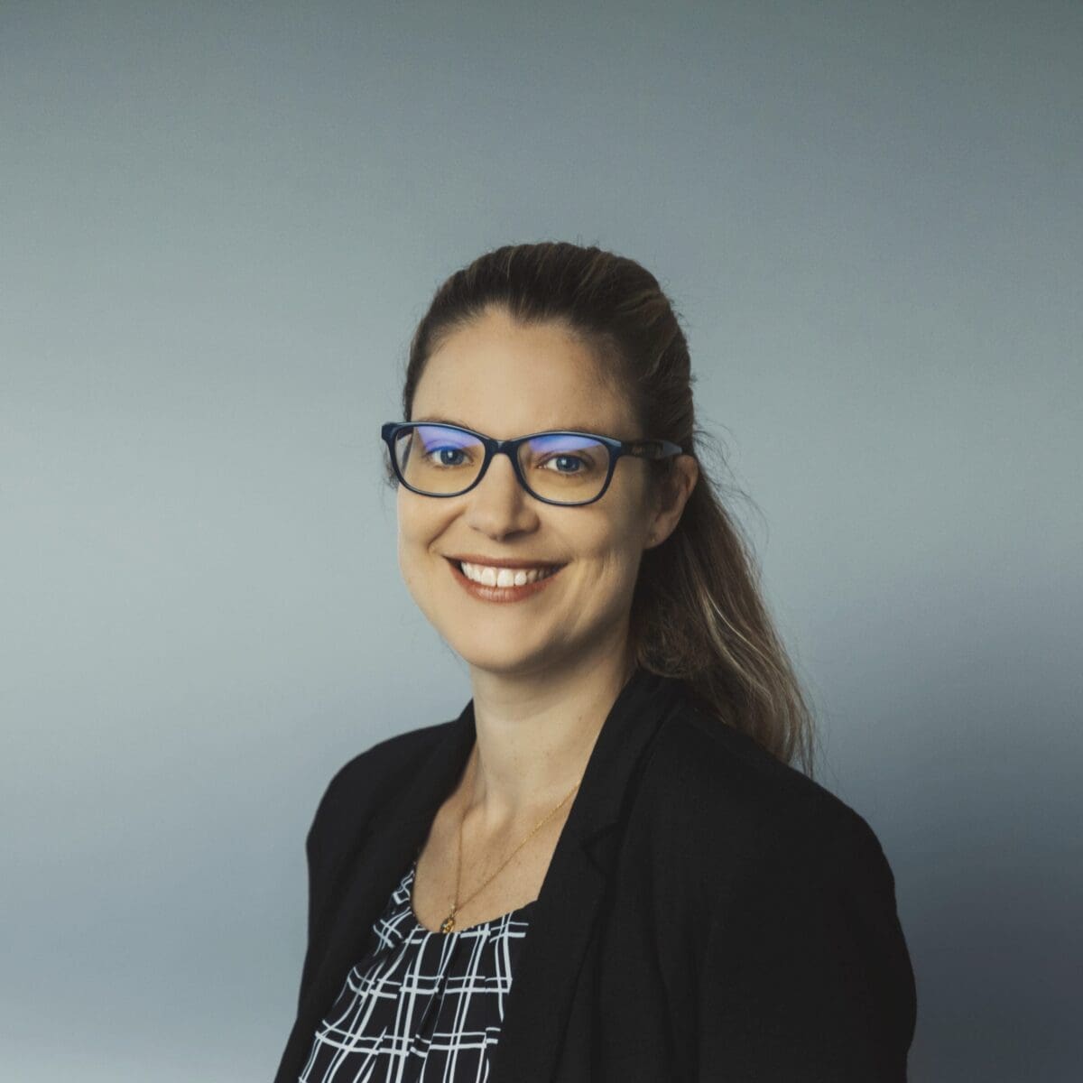 Professional headshot of a woman with glasses, black blazer, patterned top, and grey background.