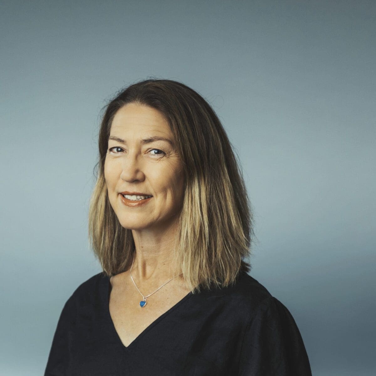 Woman with shoulder-length hair wearing a black top and blue heart pendant necklace on grey background