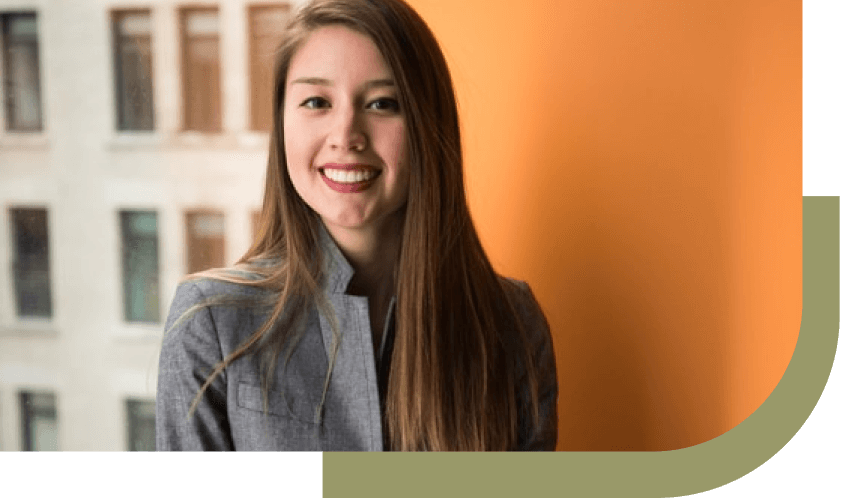 Young woman in grey blazer smiling indoors with orange wall and window view.