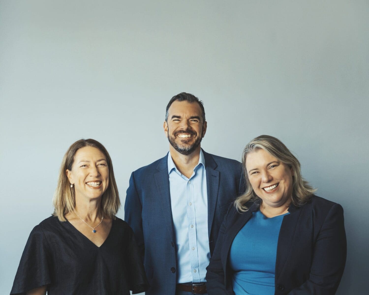 Three adults smiling in front of a plain light grey wall wearing business and casual attire.