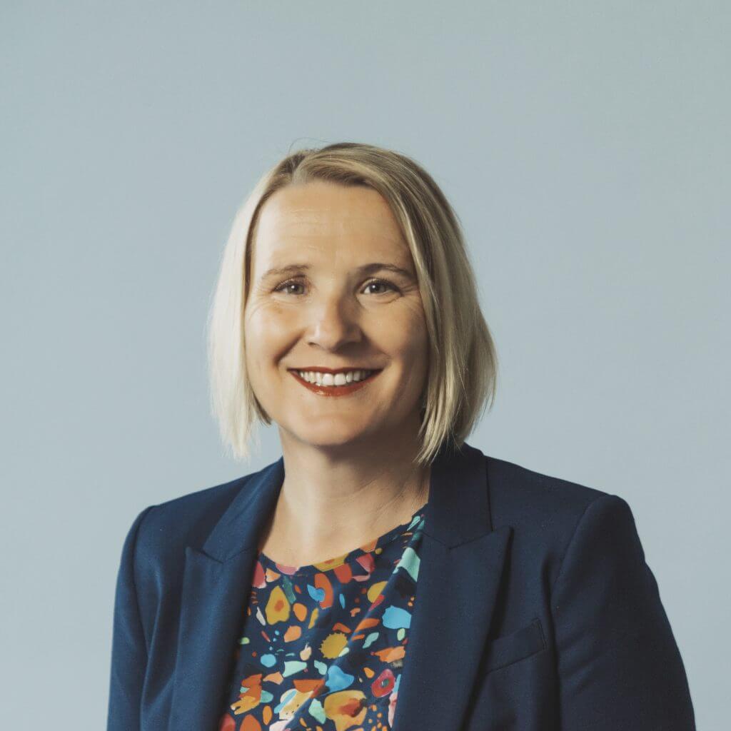 Headshot of a person with short blonde hair wearing a dark blazer and colorful patterned top.