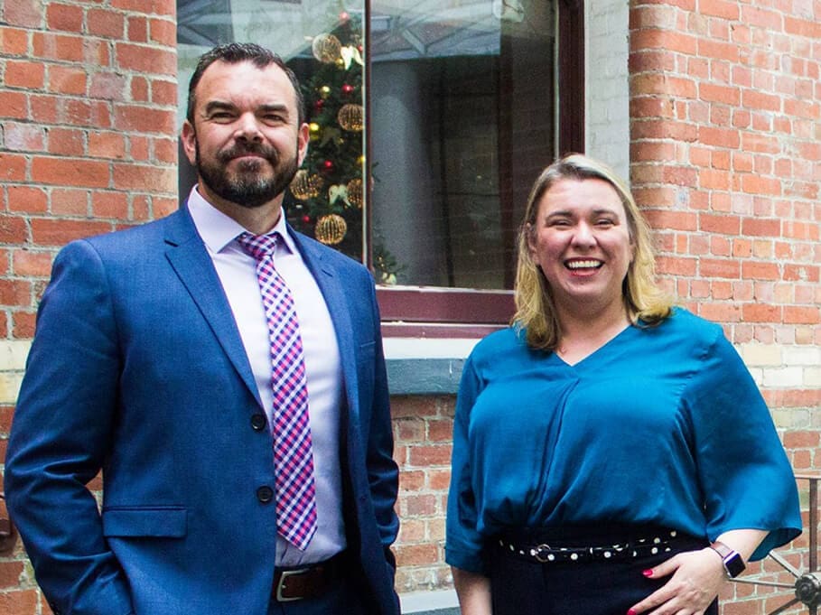 Man in blue suit and tie and woman in blue blouse stand outside brick building with Christmas tree visible.