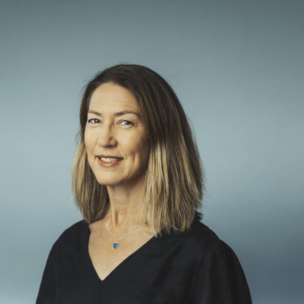 Middle-aged woman with shoulder-length light brown hair wearing black top and blue pendant necklace on grey background