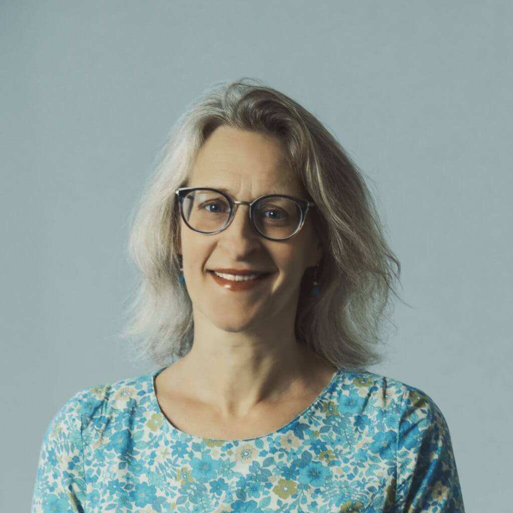 Middle-aged woman with grey hair and glasses wearing a blue floral top against grey background