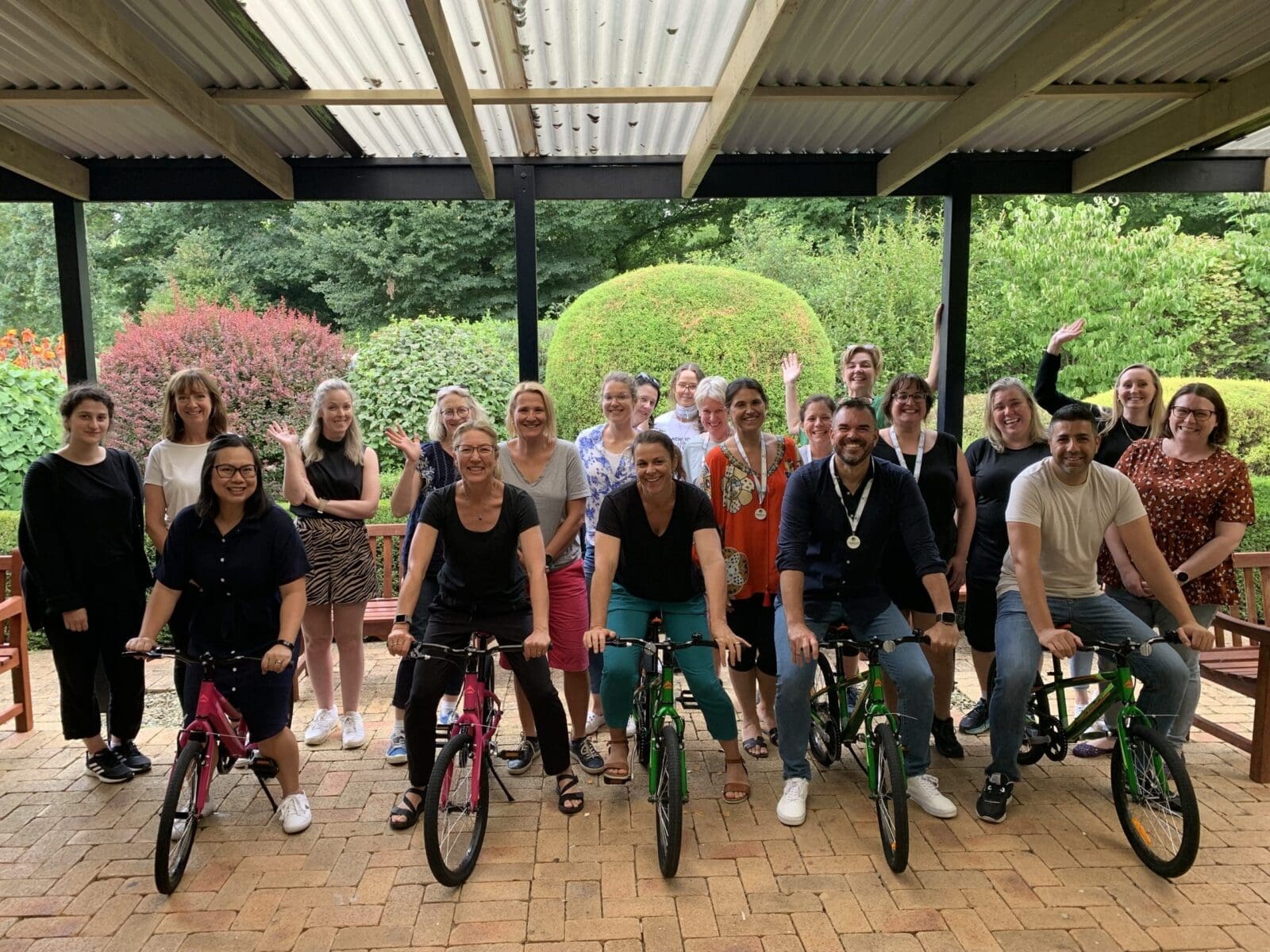 Group of 20 people, including four on bicycles, posing under a sheltered area with garden background