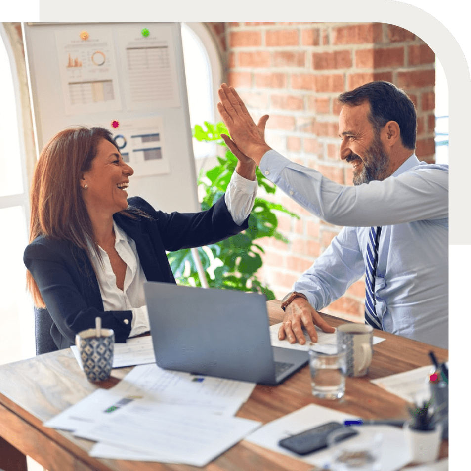 Two colleagues in business attire high five at a desk with laptop, documents, and drinks.