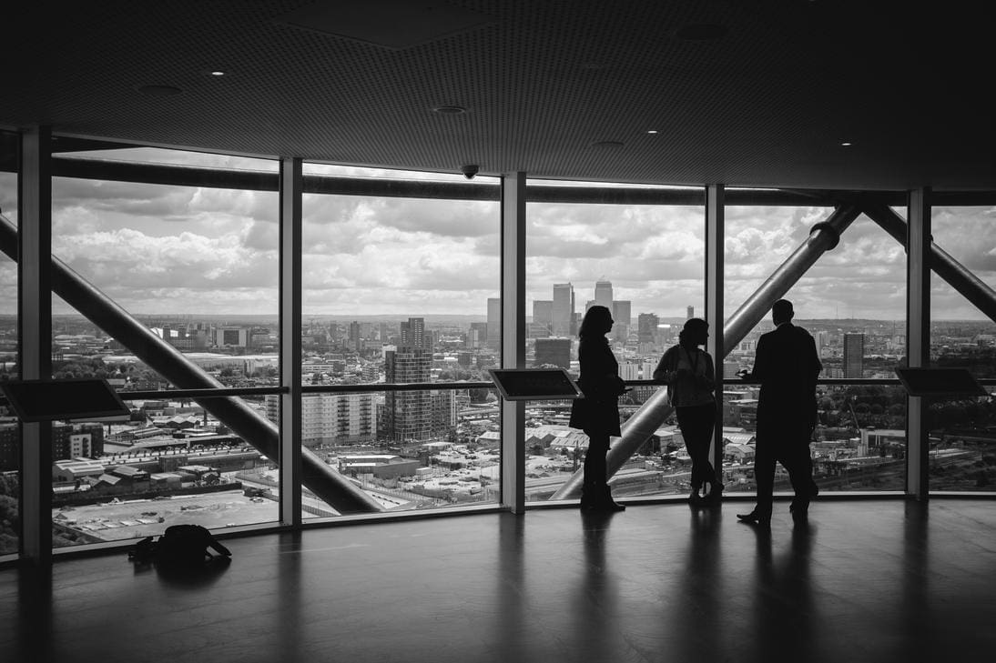 Three people stand talking inside a building with large windows overlooking a cityscape.