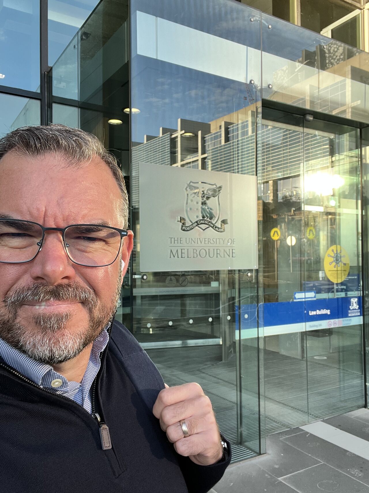 Man with glasses and beard outside University of Melbourne Law Building entrance with logo visible.